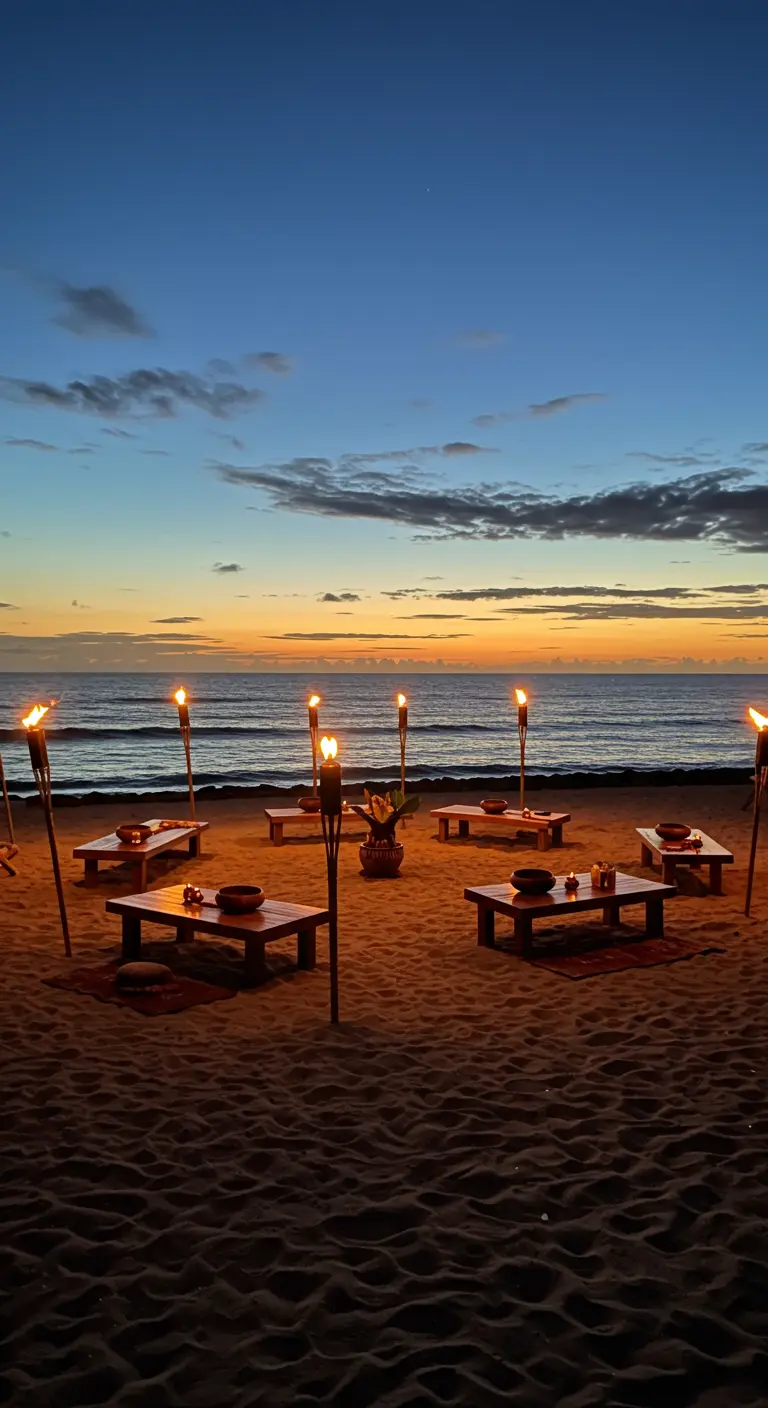Low tables set up on a beach at sunset, illuminated by tiki torches.