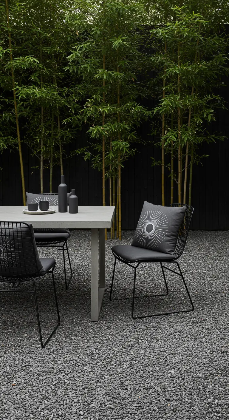 Black wireframe chairs around a concrete dining table on dark gravel, in front of a bamboo screen.