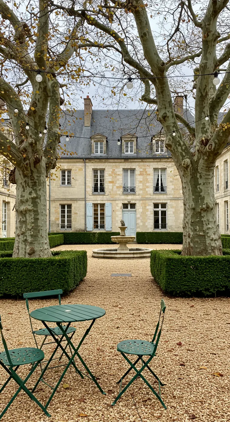 A green bistro set in a gravel courtyard between two large sycamore trees with lights.