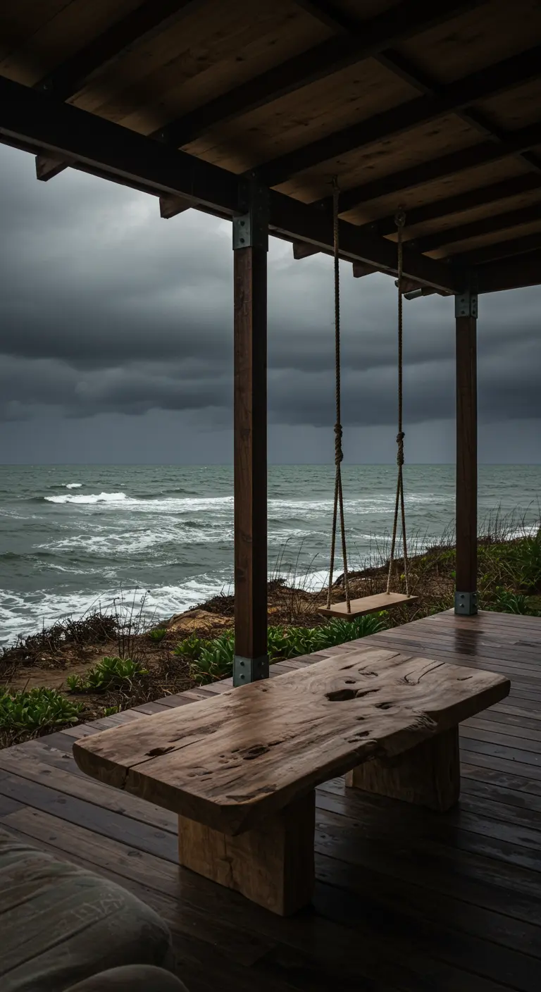 A covered porch with a dark wood table and swing overlooking a stormy ocean.