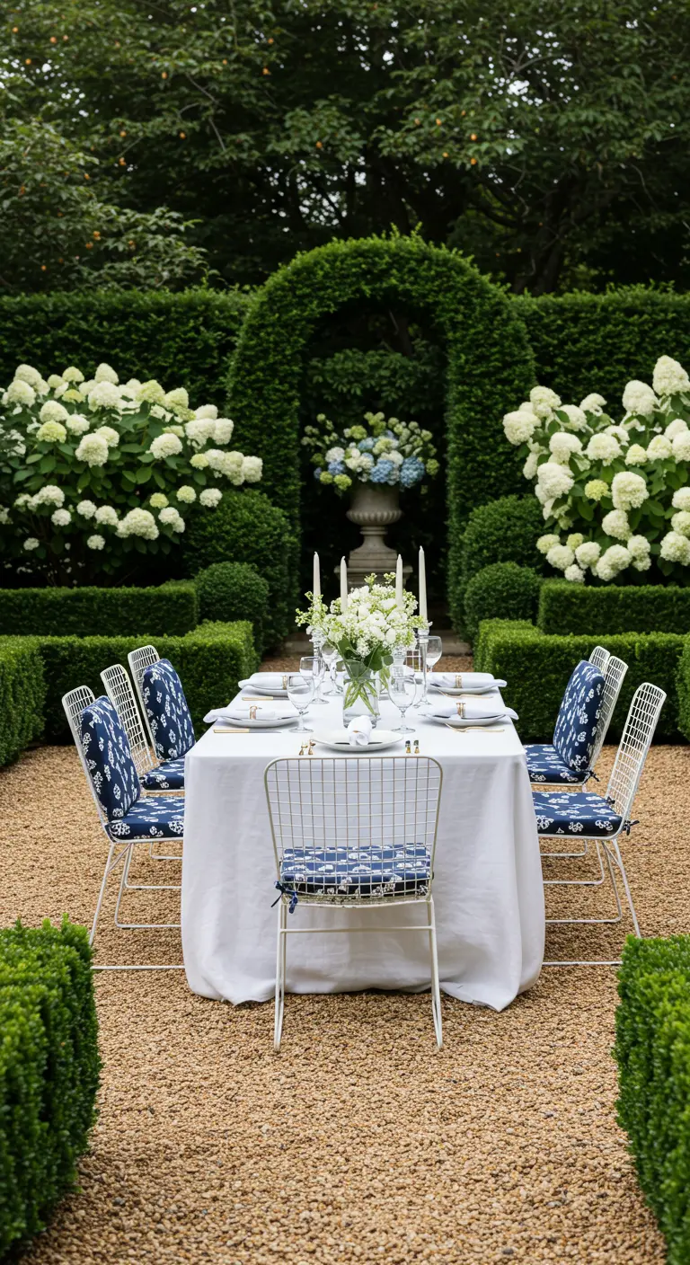 A formal garden dining setting with white wireframe chairs and blue patterned cushions around a white-clothed table.