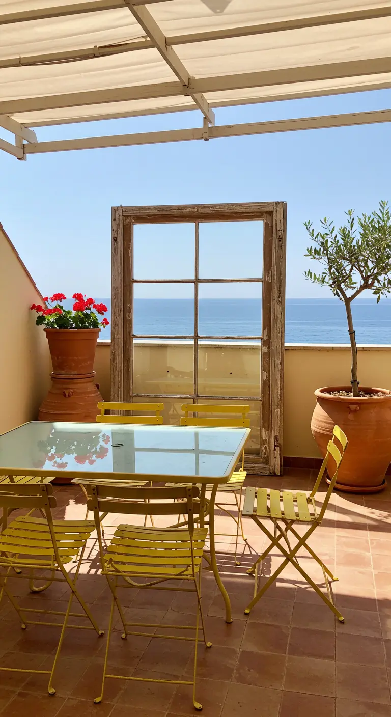 A weathered window frame on a sunny terrace frames a view of the ocean behind a yellow bistro set.