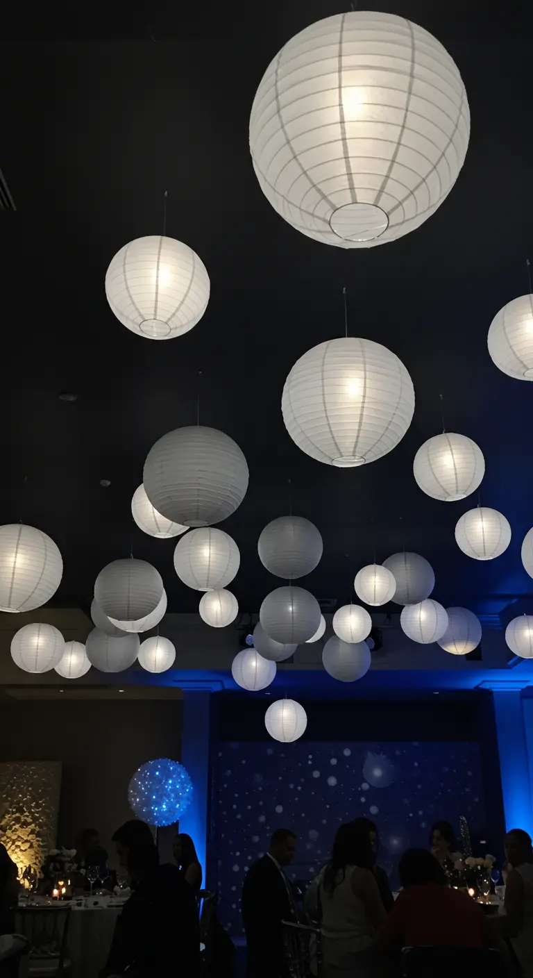 A cluster of white paper lanterns of different sizes lit from within and hanging from a dark ceiling.
