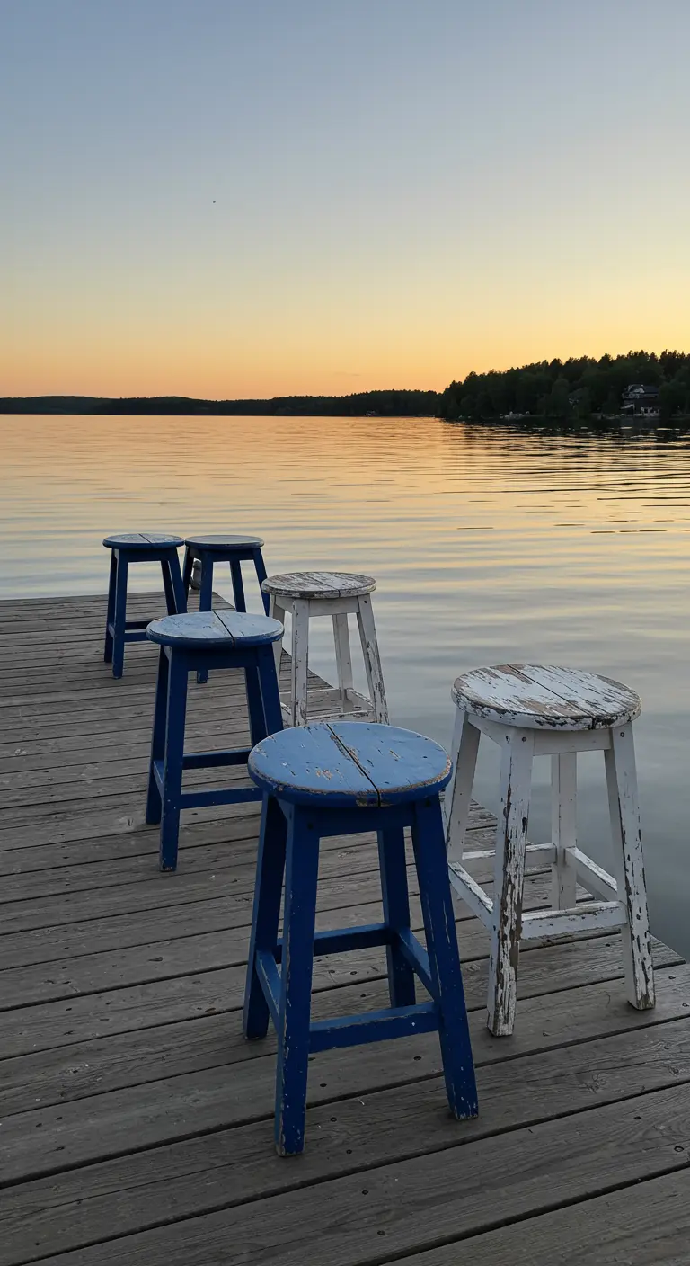 Distressed blue and white stools lined up on a wooden dock overlooking a calm lake at sunset.