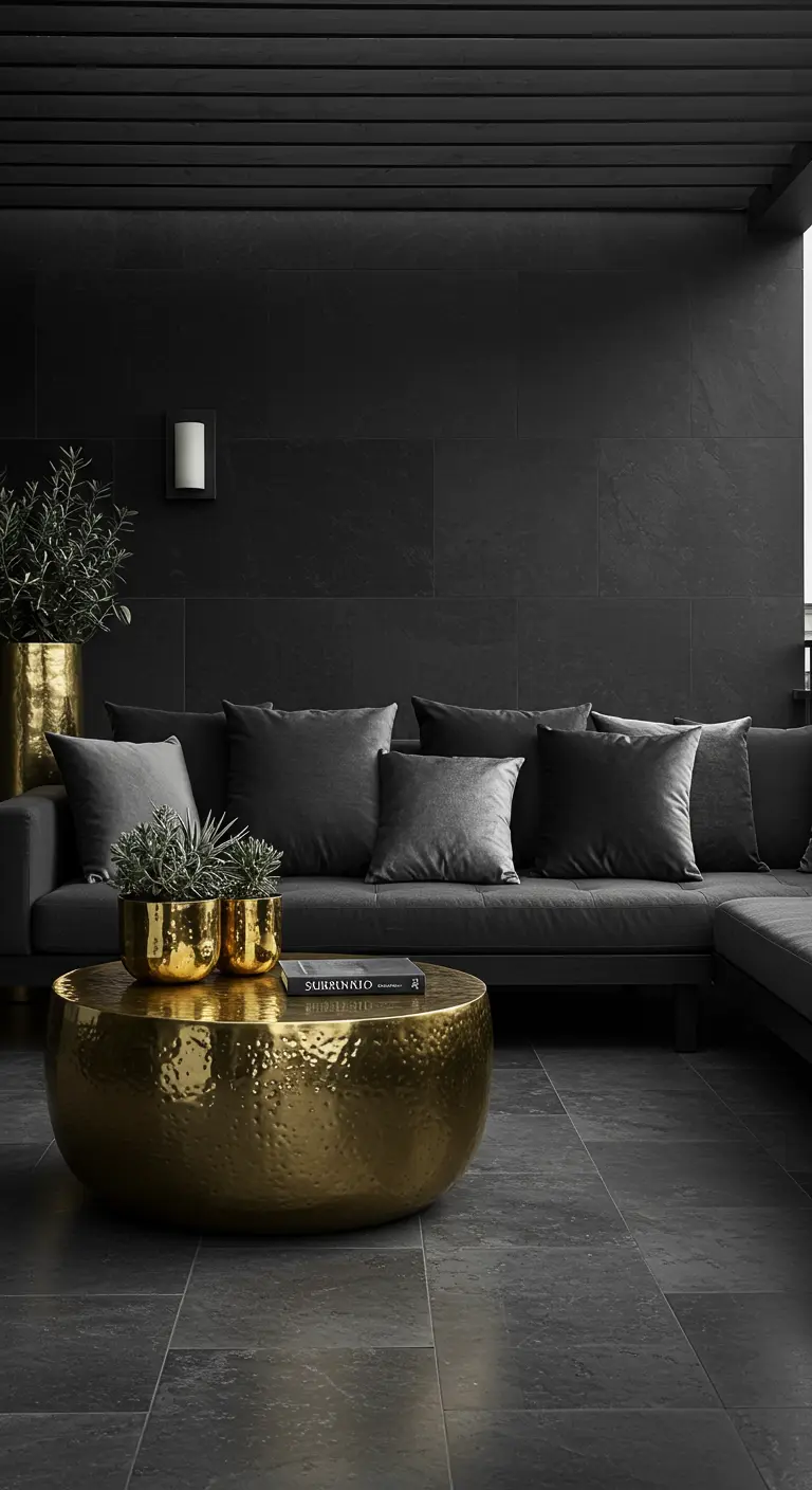 A dark monochrome lounge with a charcoal sofa, grey velvet pillows, and a large hammered gold table.
