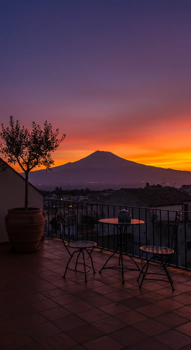 A balcony with a silhouette of an olive tree and bistro set against a fiery sunset over a volcano.