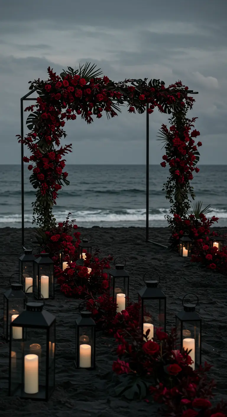 A dramatic wedding arch with red roses on a black sand beach, with a lantern-lined aisle.