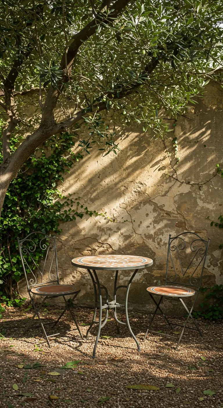 Small mosaic terra cotta bistro table with two iron chairs under an olive tree.