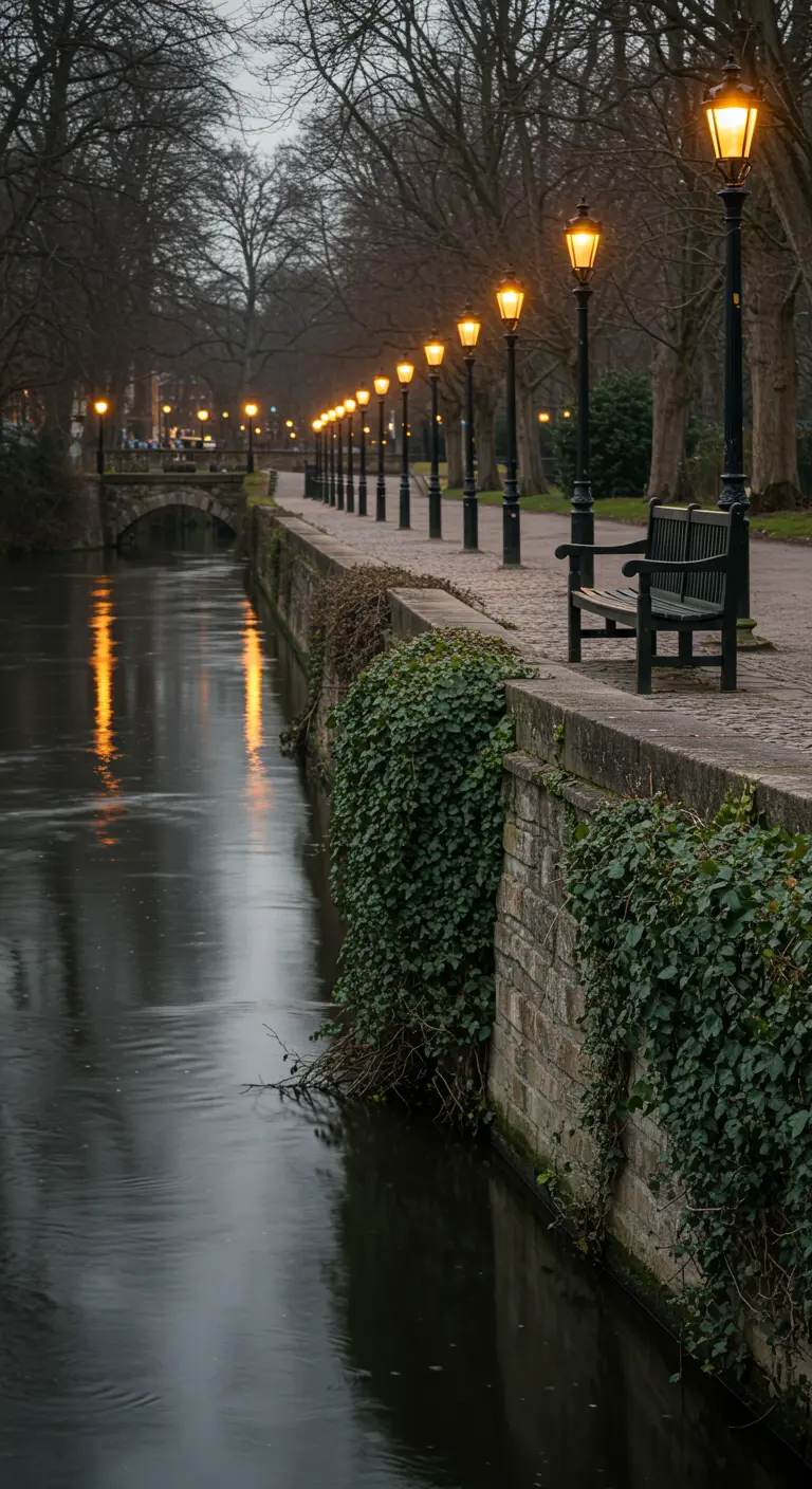 A long, straight row of lit lampposts illuminates a path and bench along a canal at dusk.