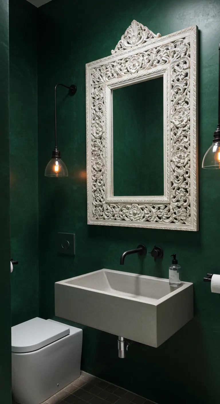 A dark green powder room with a large, ornate, whitewashed carved mirror over a concrete sink.